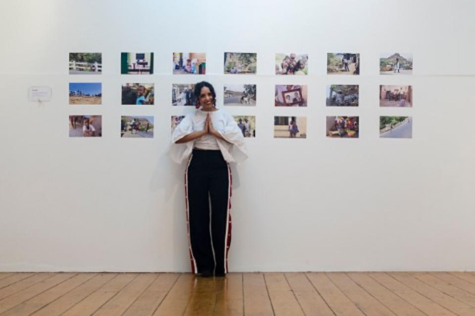 Janeth Tavares de mãos juntas em frente à parede de fotografias da exposição NÓS DE NÓS